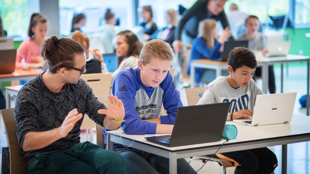 Kinderen (jongens) zitten in de klas achter een laptop. Ze krijgen les van een leraar
