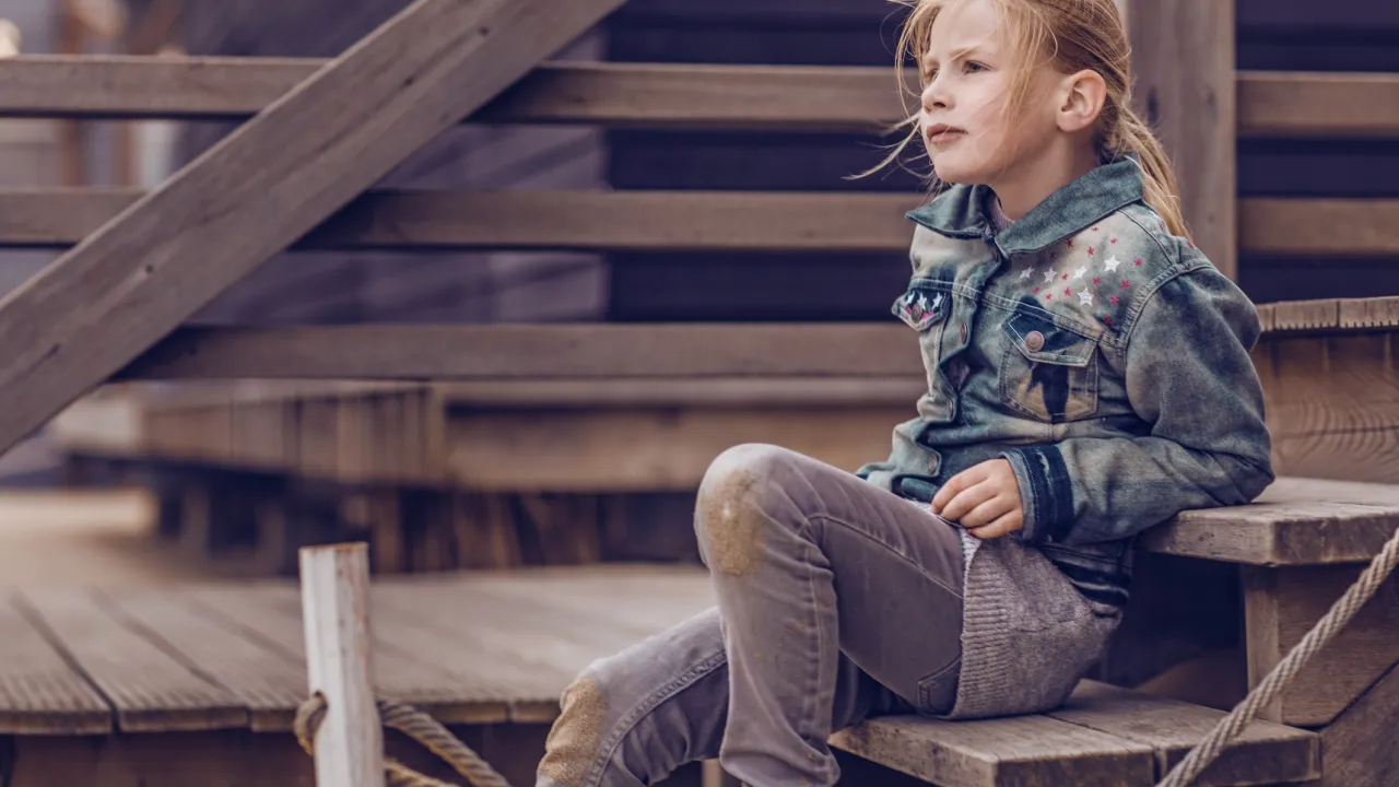 Meisje met blond haar zit op een trappetje bij een huisje op het strand. Ze kijkt voor zich uit.