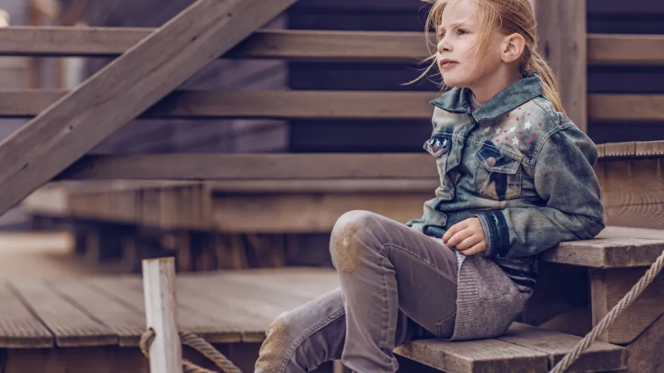 Meisje met blond haar zit op een trappetje bij een huisje op het strand. Ze kijkt voor zich uit.