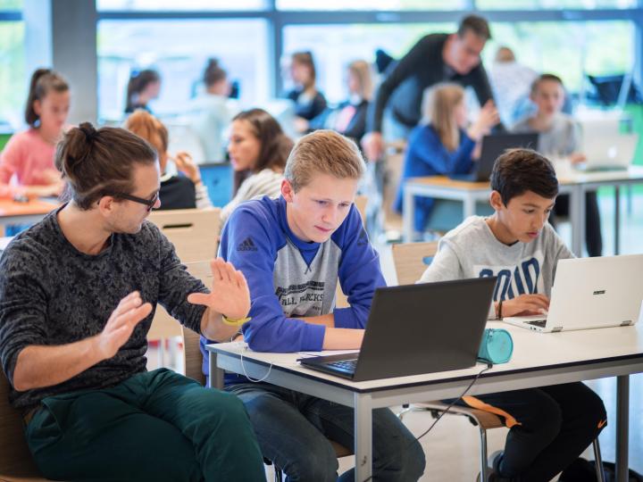 Kinderen (jongens) zitten in de klas achter een laptop. Ze krijgen les van een leraar