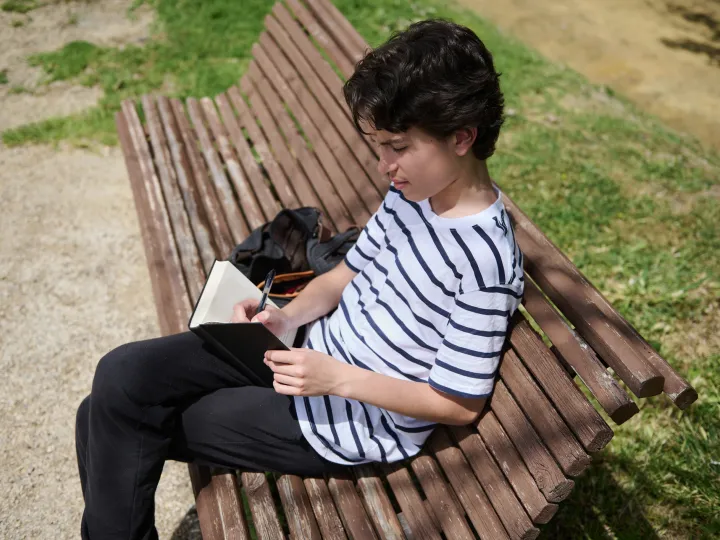 Jongen zit op bankje buiten en schrijft in boekje